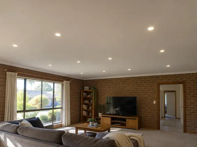 Living room with recessed downlights, brick walls, large window and TV on a beige carpet in Wyong