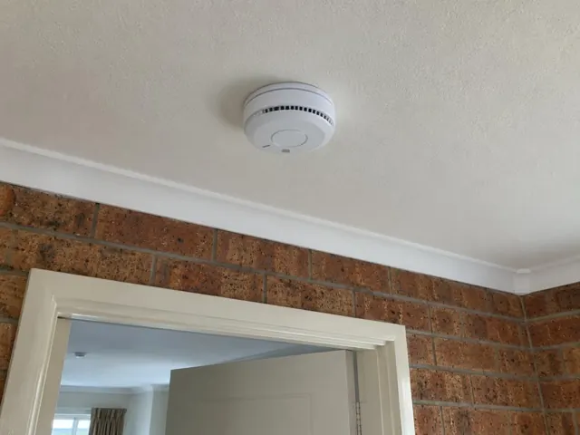 Ceiling smoke alarm above a doorway in a brick-walled home interior in Tumbi Umbi